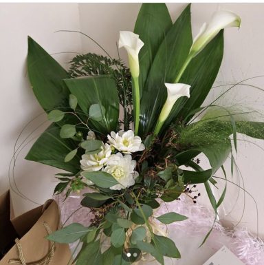 A floral arrangement featuring white calla lilies, green foliage, and delicate flowers.