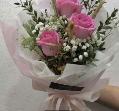 Bouquet of pink roses with white flowers and greenery, wrapped in delicate paper.